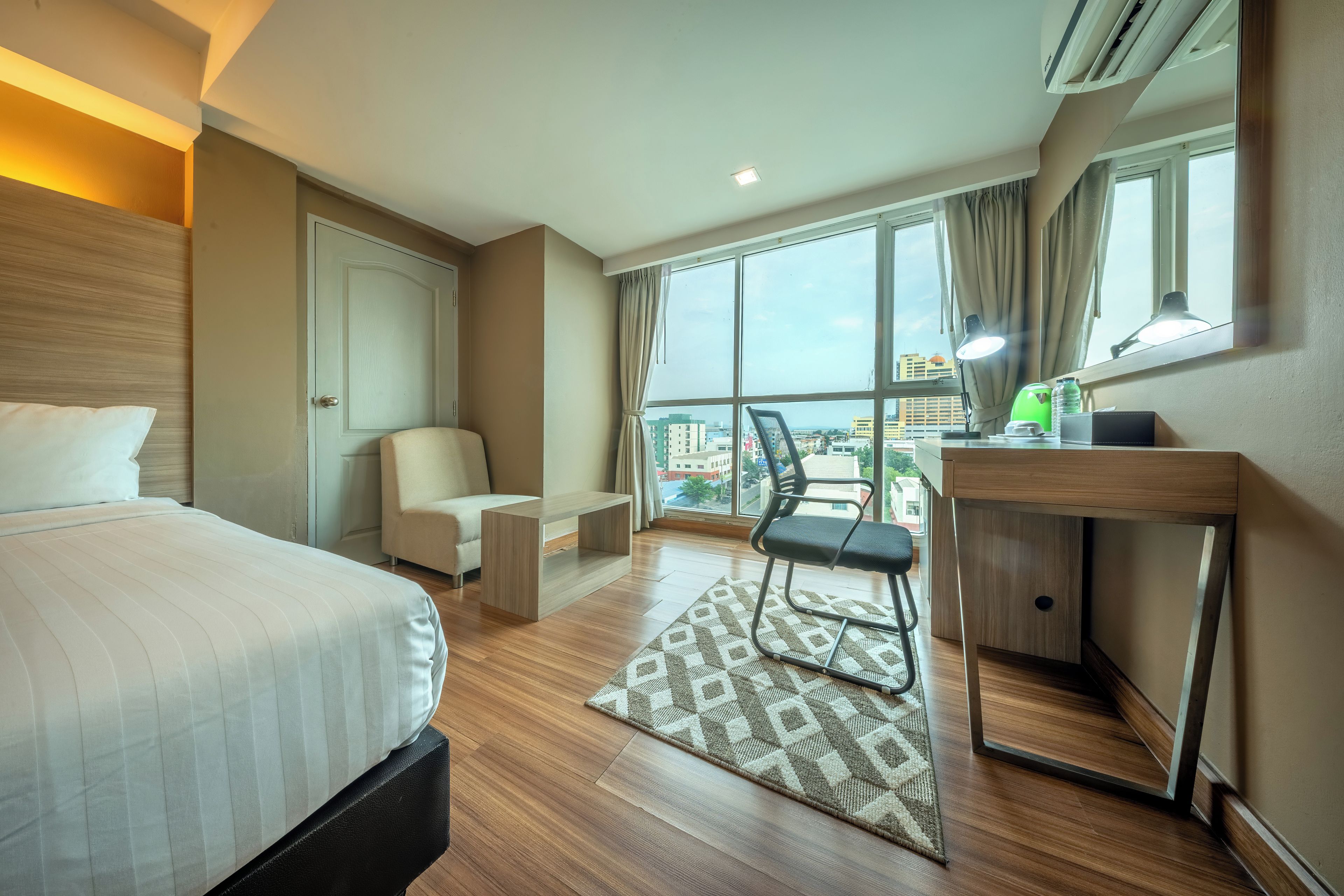 Habitación de hotel con cama, escritorio, silla, mesita, ventana con vista a edificios y equipo de aire acondicionado.