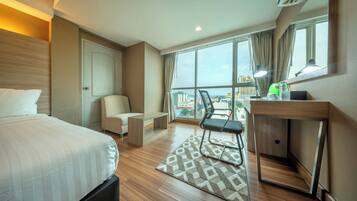 Habitación de hotel con cama, escritorio, silla, mesita, ventana con vista a edificios y equipo de aire acondicionado.