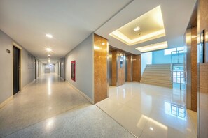 Un pasillo moderno de hotel con un ascensor central, una escalera y una entrada de vidrio.