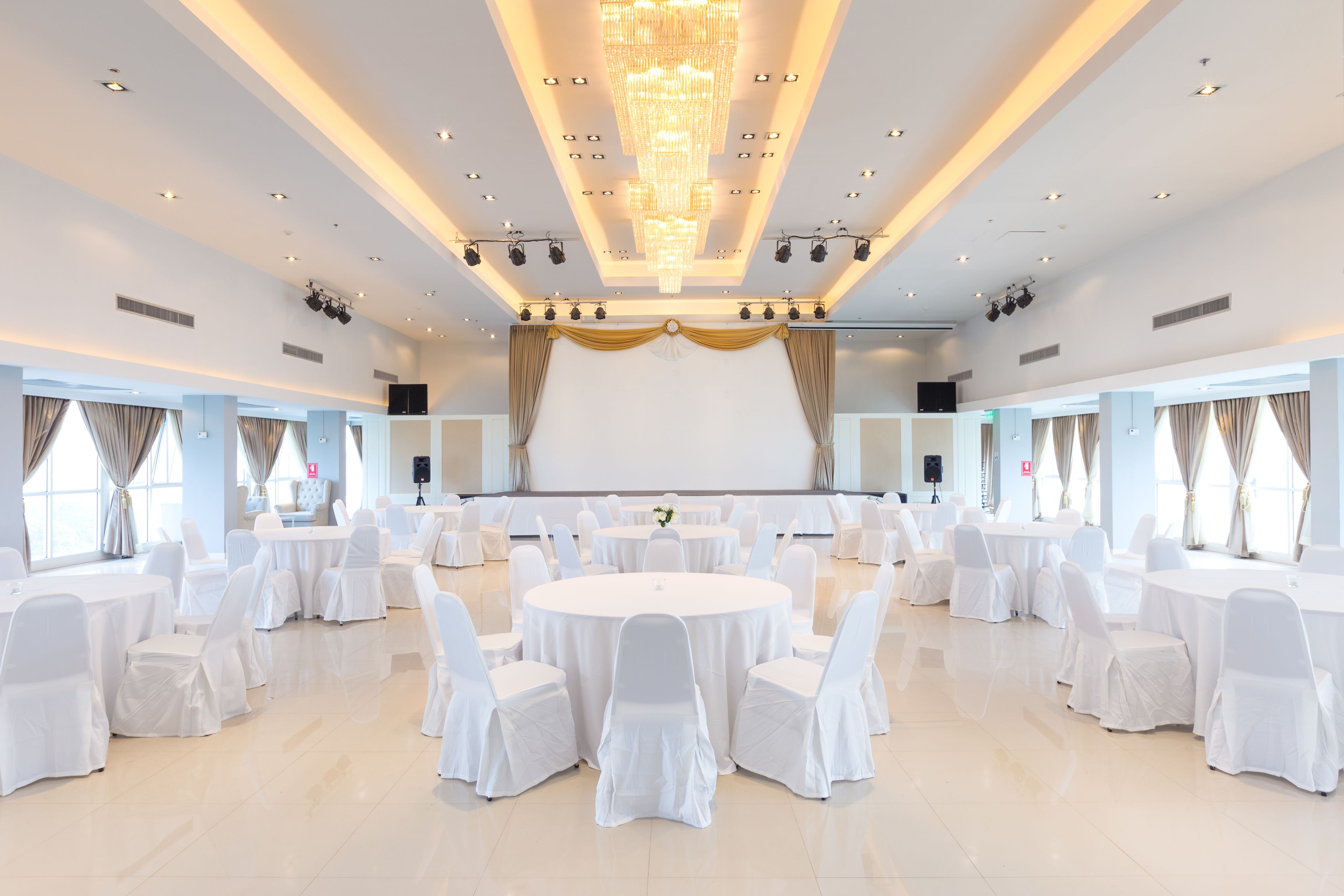 A large, well-lit banquet hall with round tables and white chairs, a stage with curtains, and a chandelier.