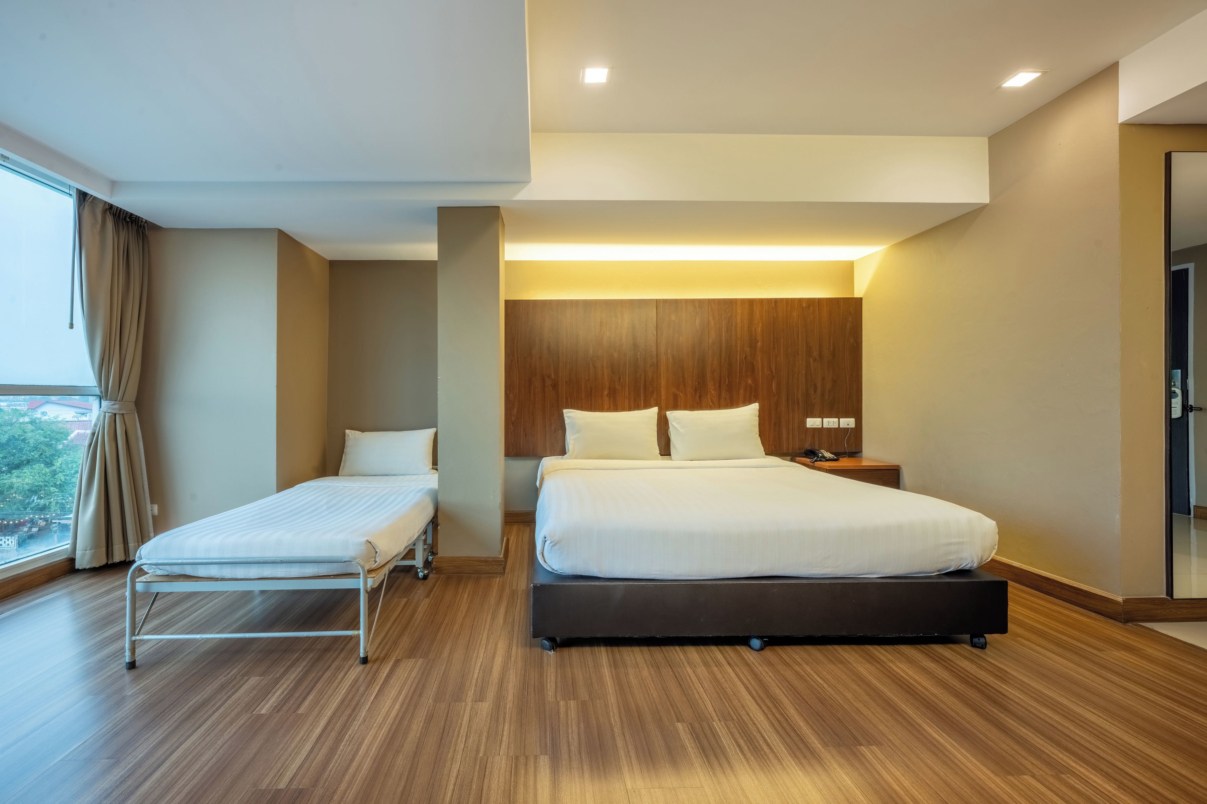 Habitación de hotel con una cama grande, una cama más pequeña, cabecera de madera, mesita de noche y vistas al exterior.