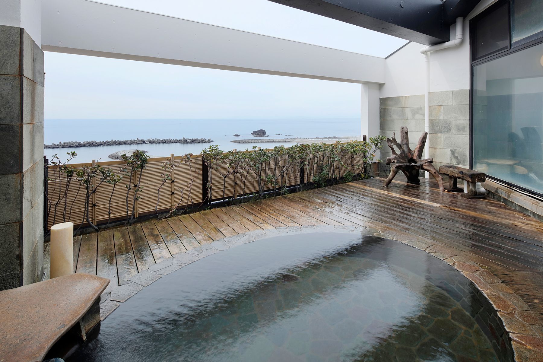 Une terrasse en bois avec une piscine bordée de pierres, offrant une vue sur une mer calme parsemée de rochers.