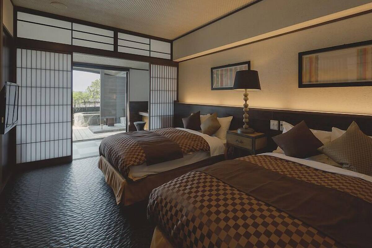 A hotel room with two beds, a sliding glass door to a balcony, a bedside table with a lamp, and a framed picture on the wall.