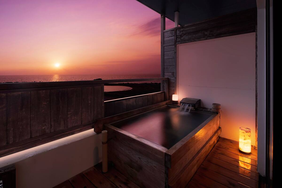 A wooden deck with a hot tub and a lantern, overlooking a sunset over the ocean.