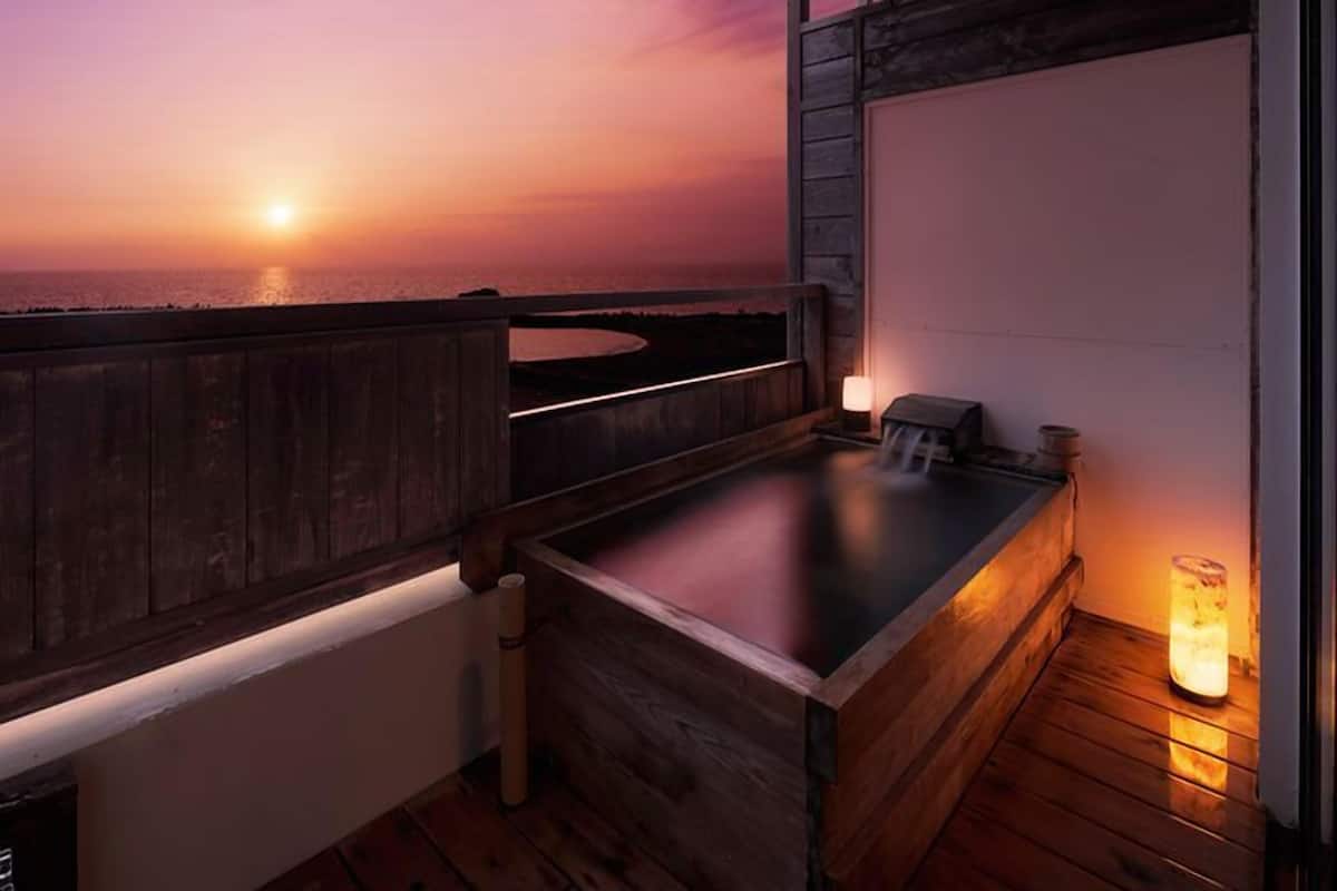 A wooden deck with a hot tub, a view of the sunset over the ocean, and a night lamp.
