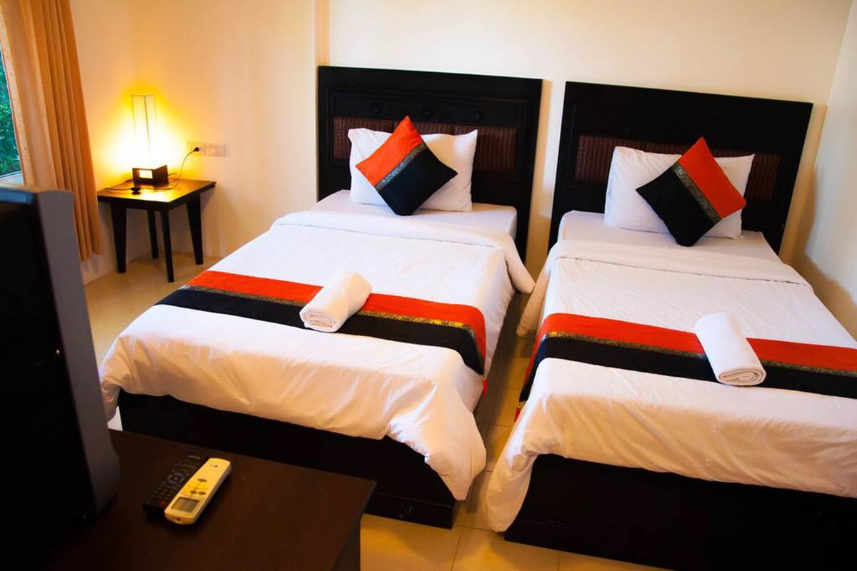 Habitación de hotel con dos camas, cada una con ropa de cama blanca y detalles en rojo y negro. Hay una mesita de noche con una lámpara y un control remoto en el suelo.