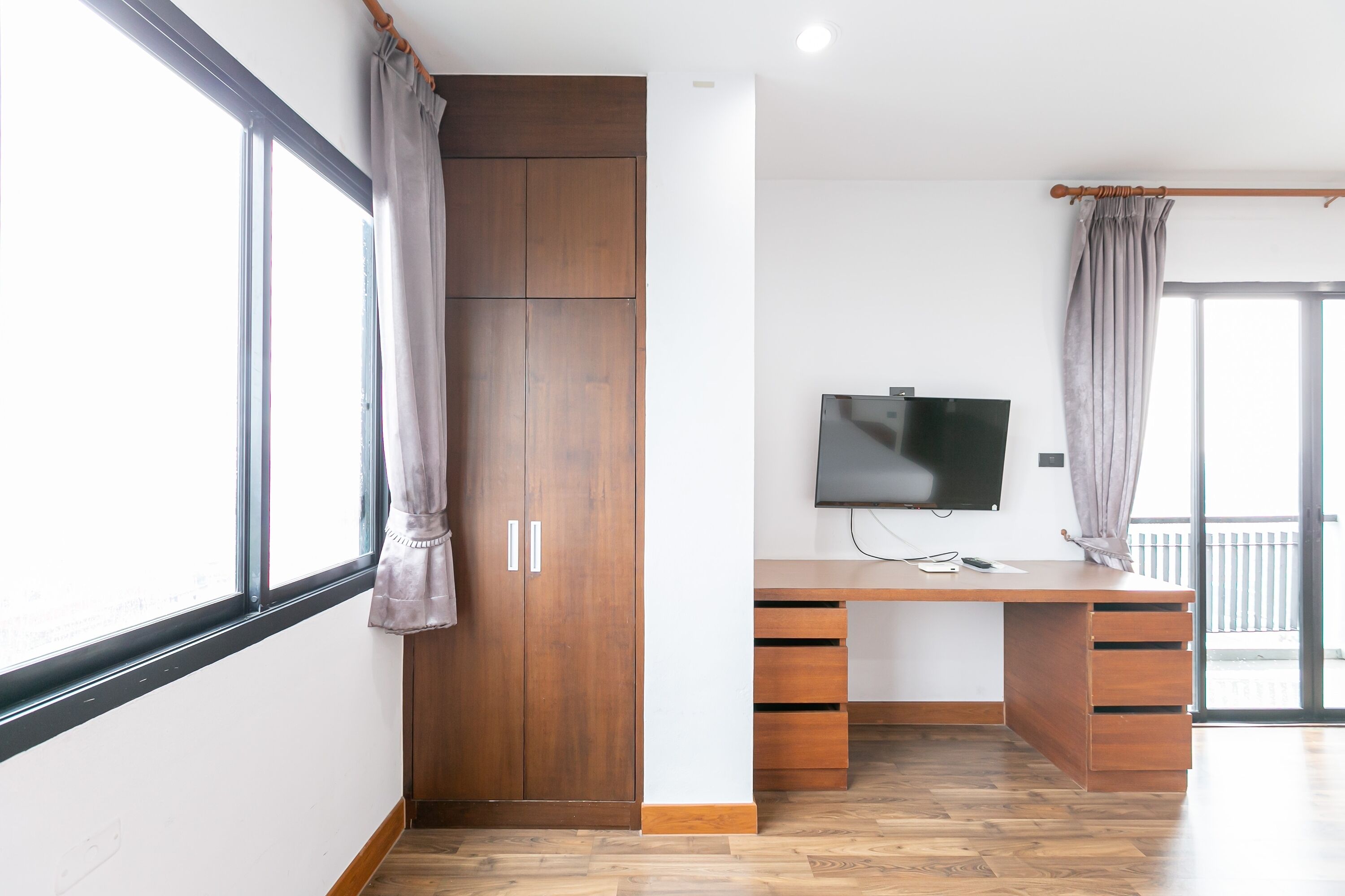 Una habitación con un armario de madera, un escritorio con cajones, un televisor de pantalla plana y ventanas amplias con cortinas.