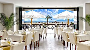 A restaurant with tables set for dining, white umbrellas, and a view of the beach. - Ayvalik Cinar Hotel (Ayvalik)