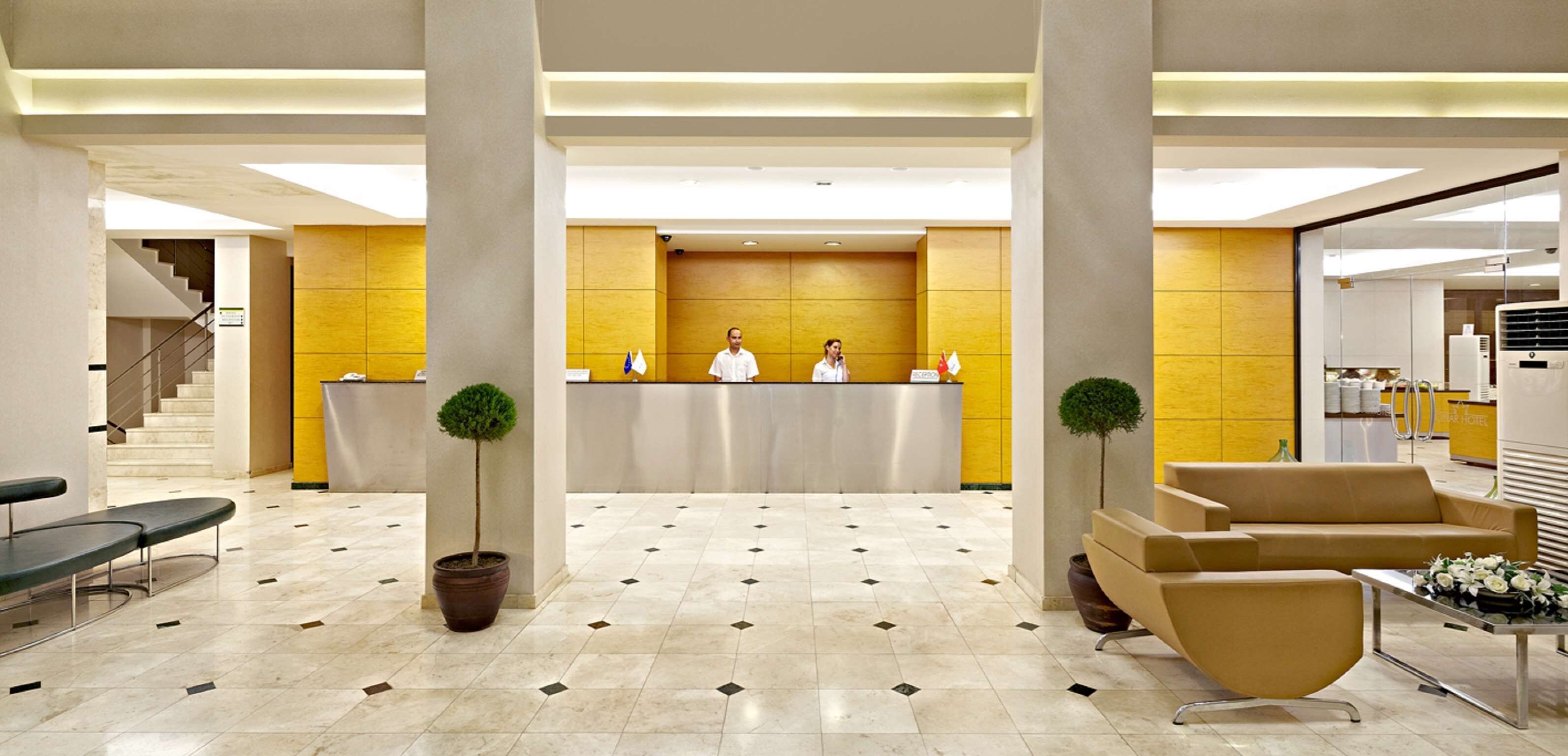 Un vestíbulo de hotel con una recepción, dos plantas en macetas, una escalera y una zona de estar.
