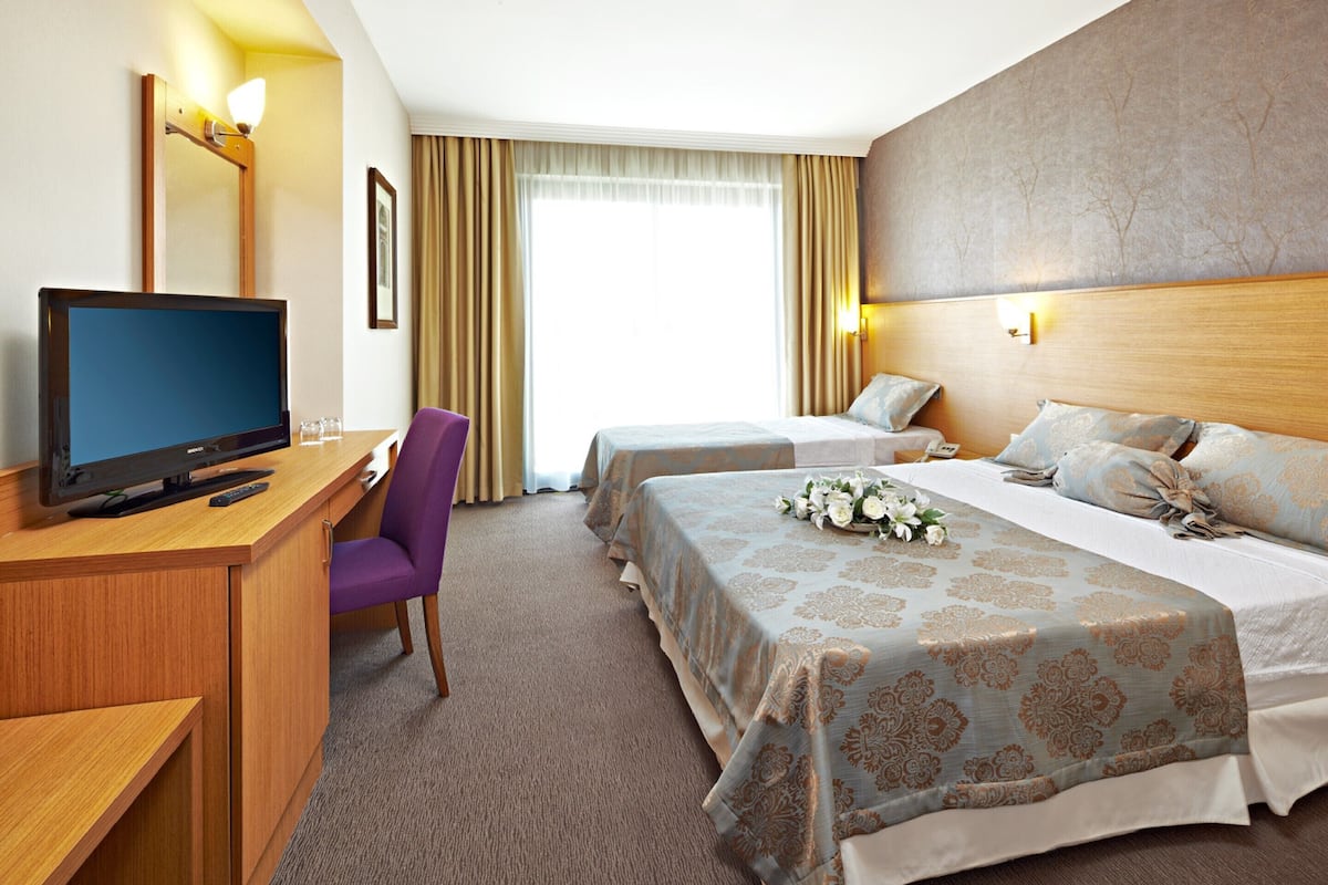 A hotel room with a TV, desk, purple chair, two beds, and a large window with curtains.