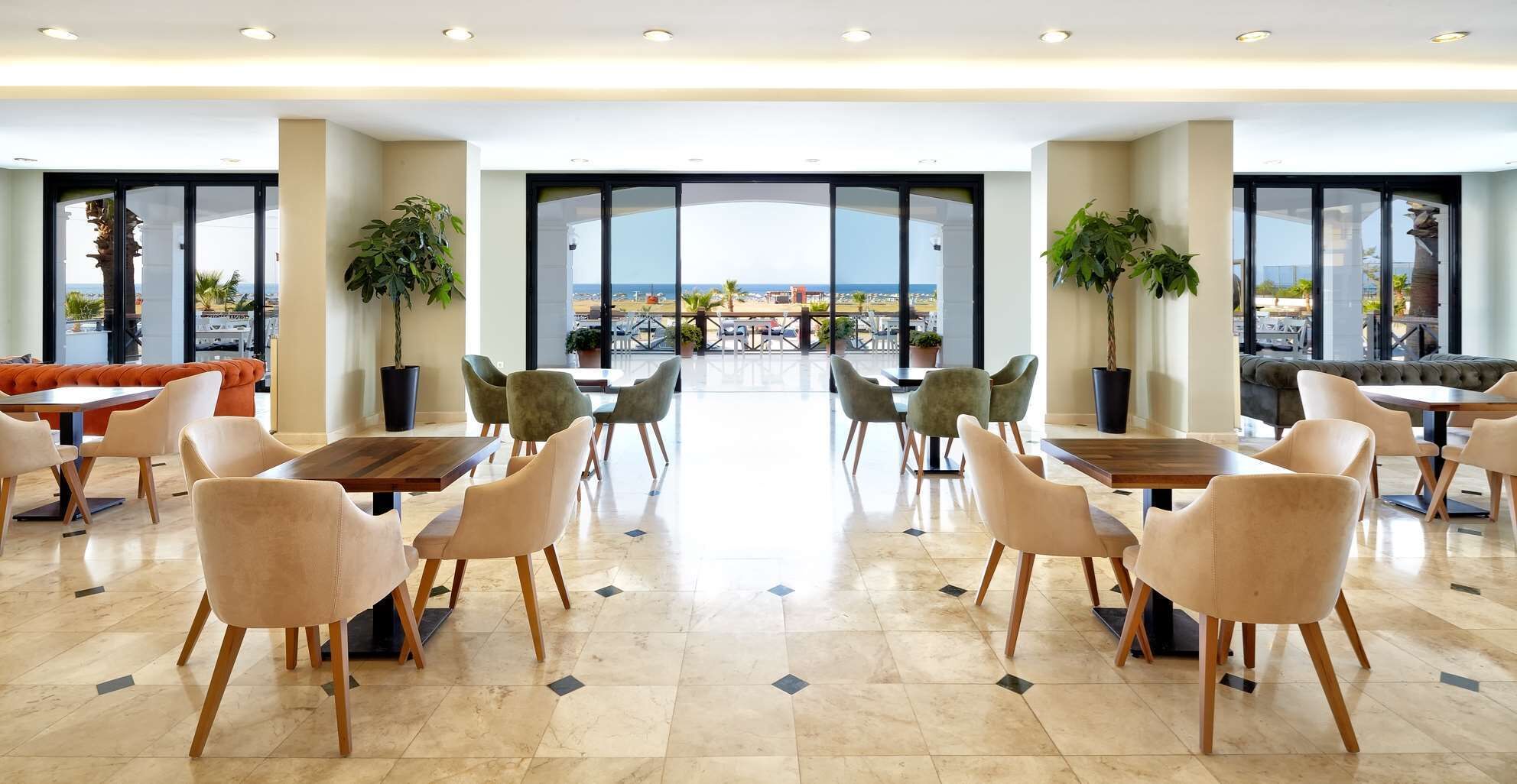 Un espace intérieur vaste et lumineux, aménagé avec différents coins salon, de grandes fenêtres donnant sur la mer et des plantes en pot.