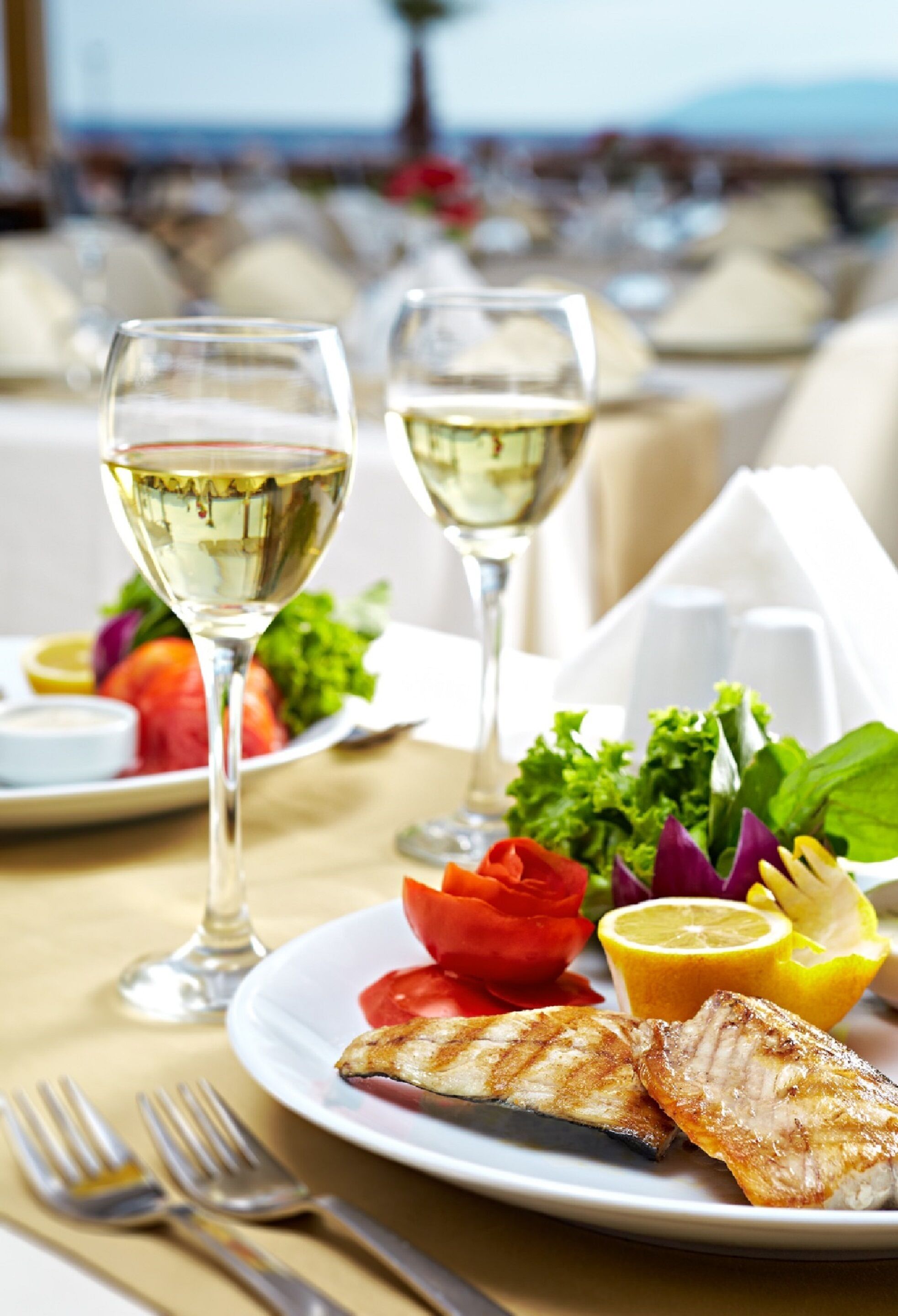Une table à manger avec une assiette de poisson grillé, une salade et deux verres de vin blanc.