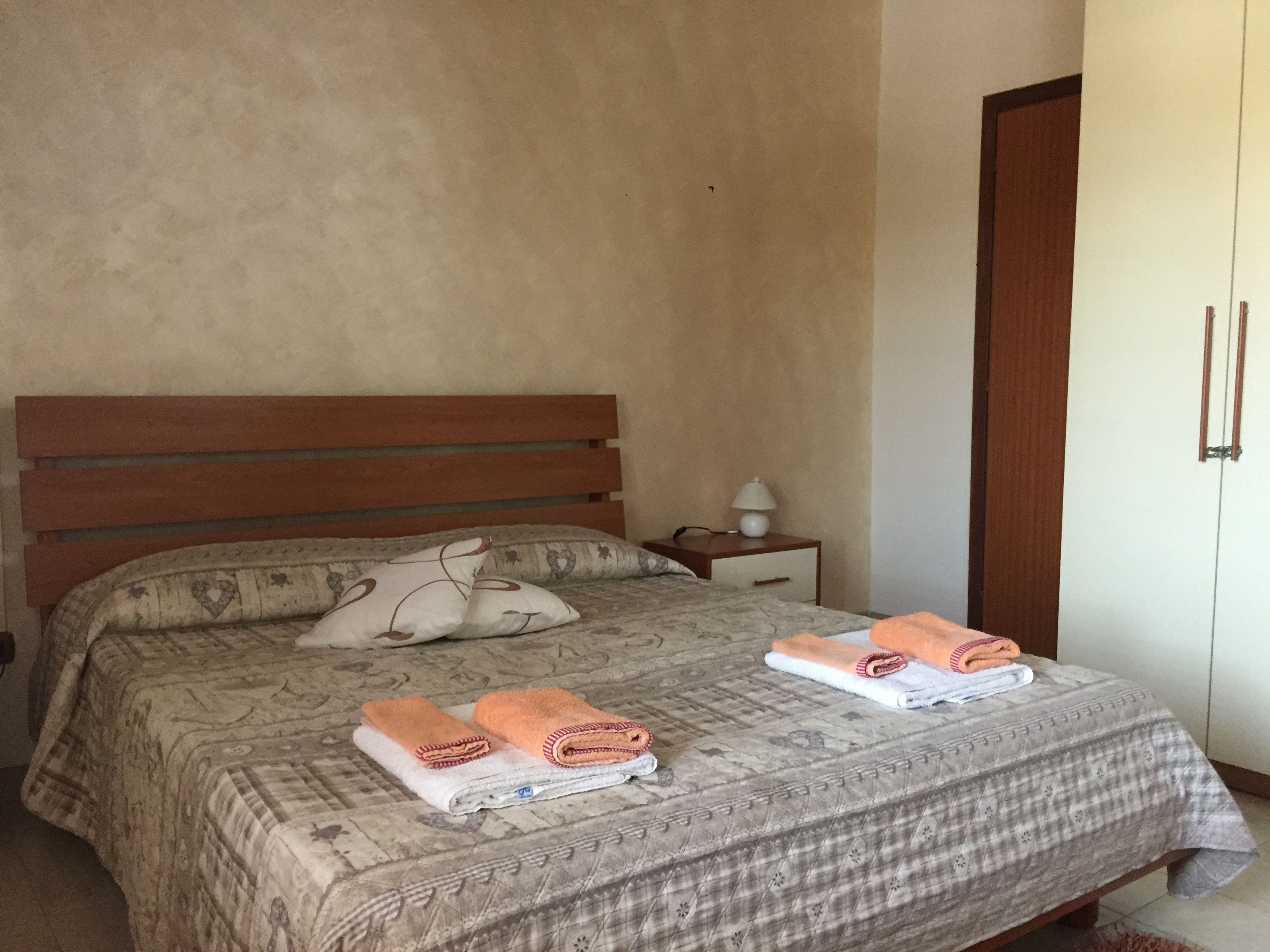 Quarto com cabeceira de madeira, cama com roupa de cama estampada, duas toalhas enroladas, mesa de cabeceira com abajur e guarda-roupa.