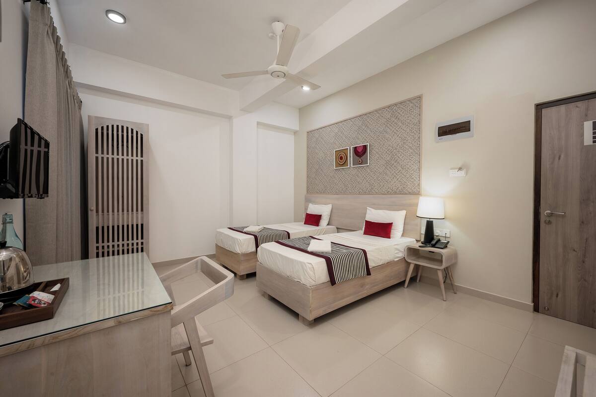 Une chambre d’hôtel avec deux lits, un bureau, une chaise, un ventilateur de plafond, un téléviseur fixé au mur et une table de chevet avec une lampe.