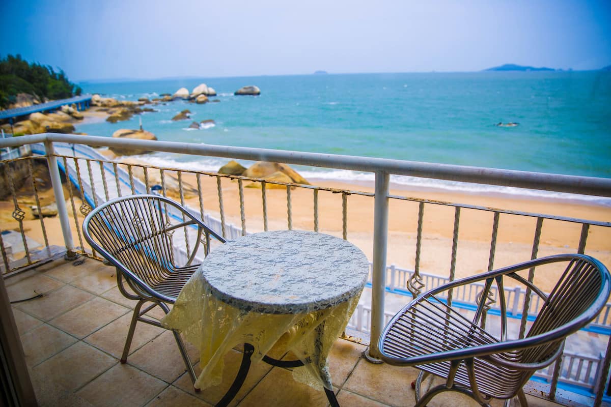 A balcony with a table and two chairs overlooking a beach with rocks and the sea.