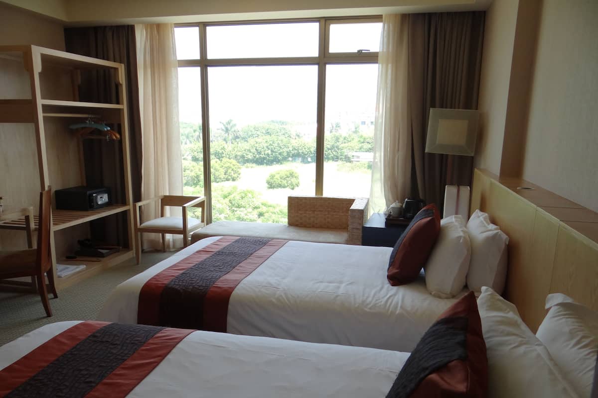 A hotel room with two beds, a desk, a chair, and a window with a view of greenery.