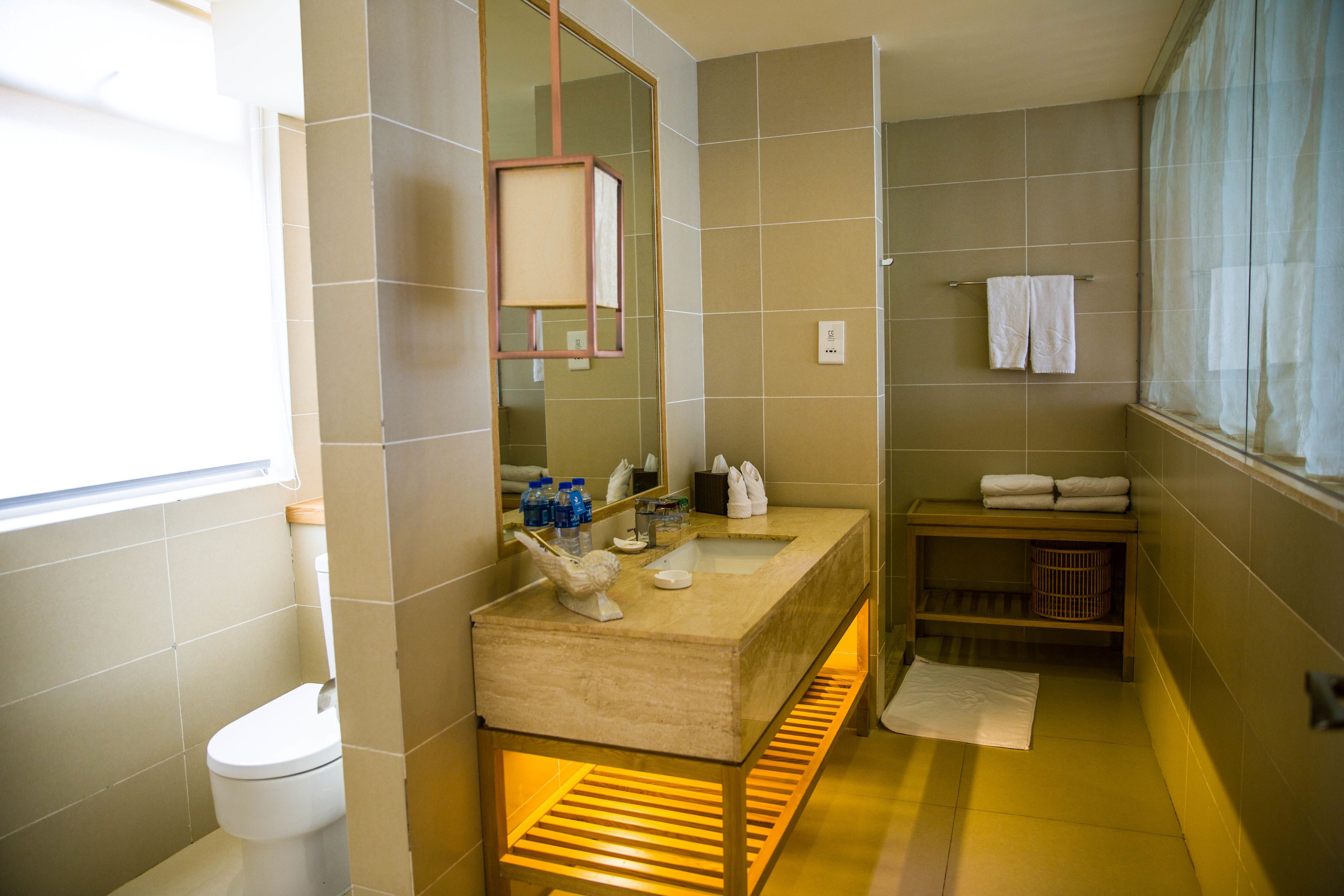 A modern bathroom with a beige tiled wall, a wooden vanity, a large mirror, and a window with curtains.