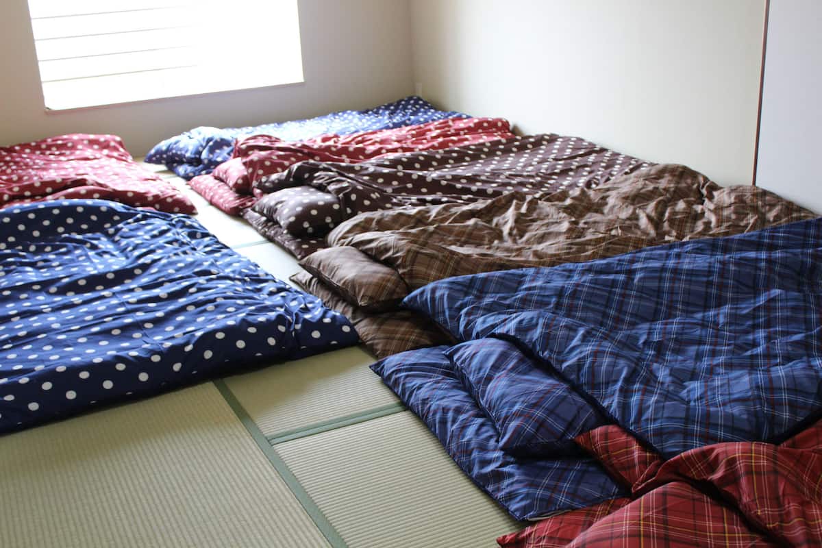 A bed with multiple blankets and pillows, a window with blinds, and a wall with a switchboard.