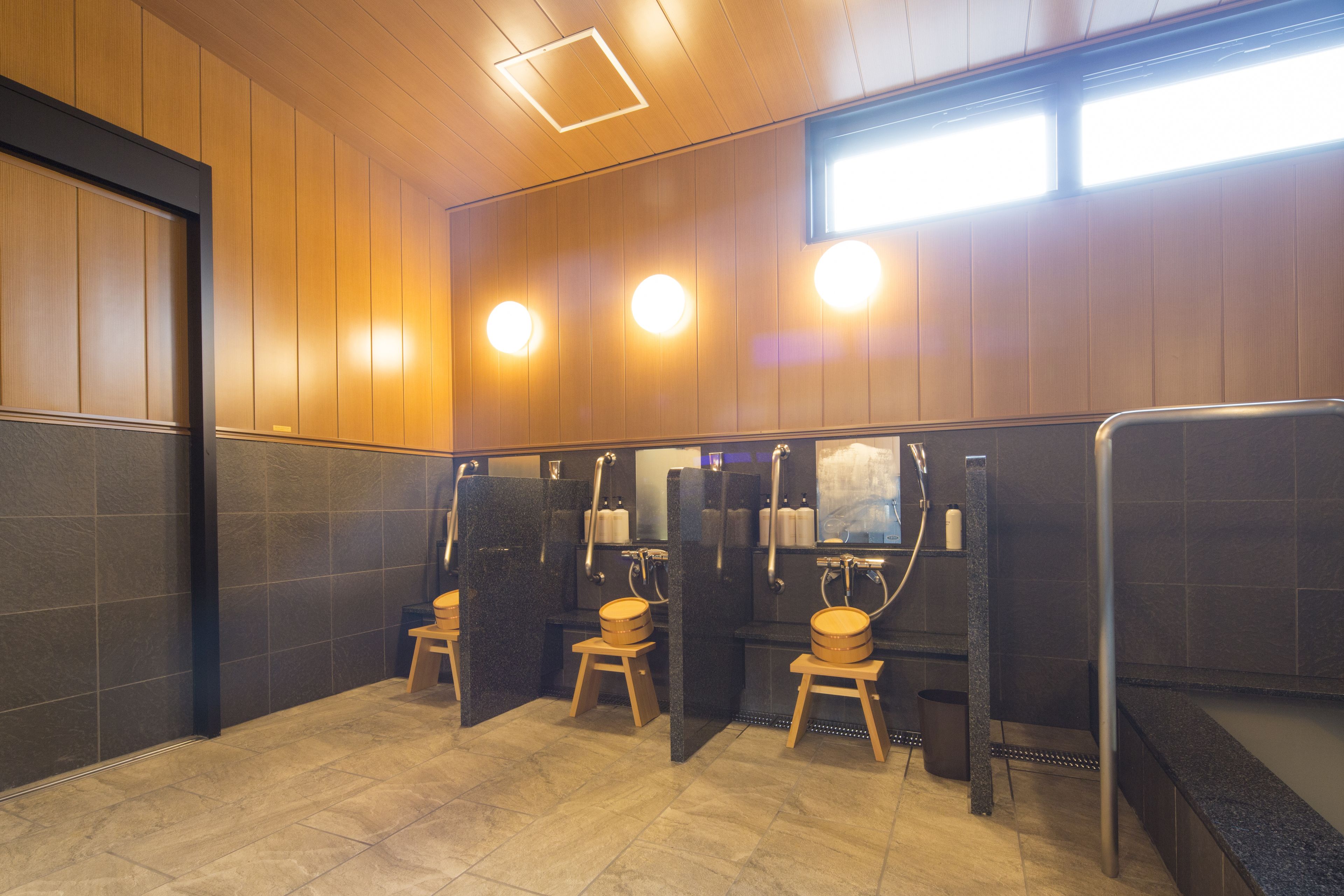 Baño público con paredes de paneles de madera, varios lavamanos y taburetes individuales.