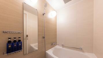 A modern bathroom with a bathtub, a shower, and a mirror. There are three bottles of shampoo and conditioner on a shelf.