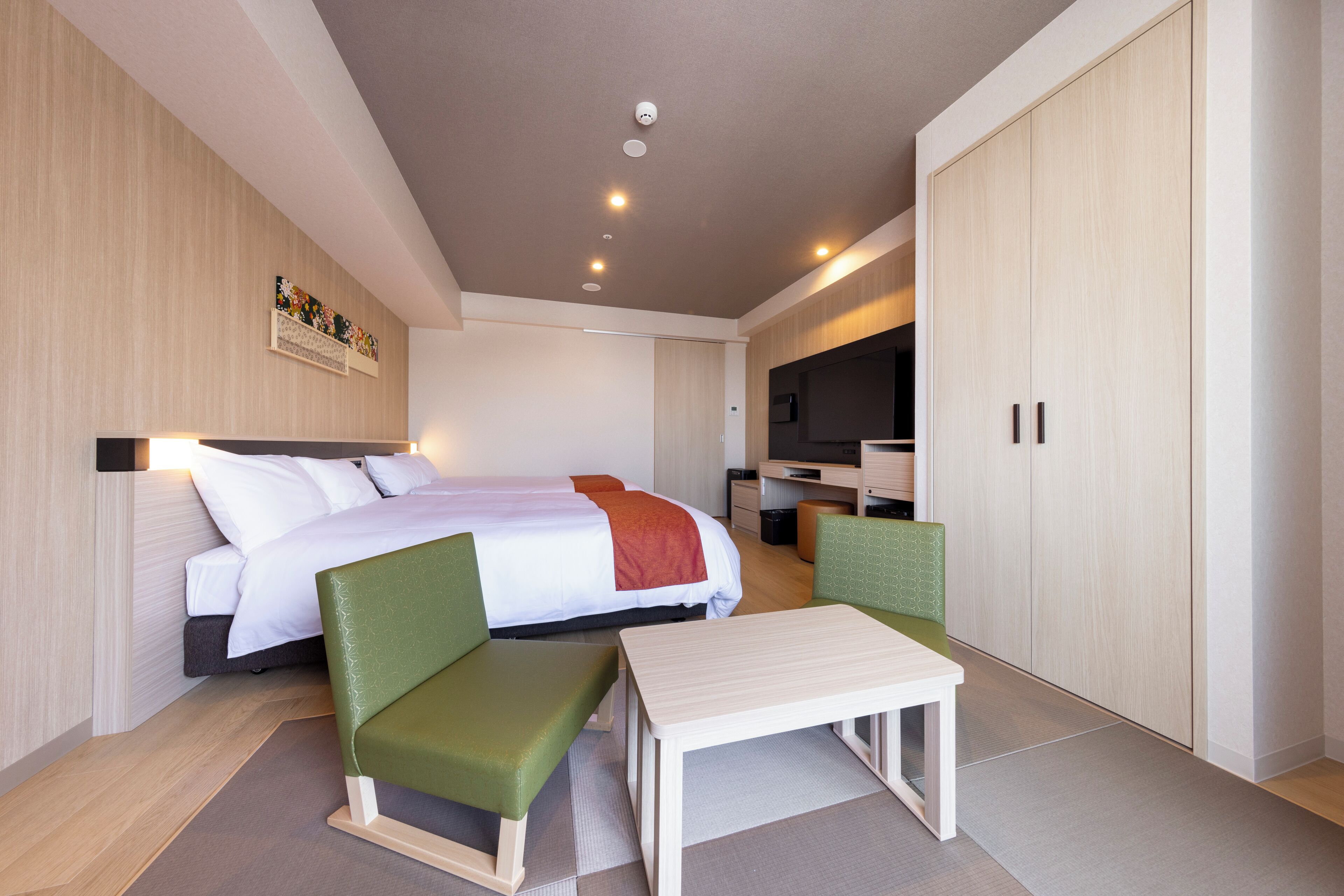 Una habitación de hotel con una cama, dos sillas verdes, una mesita, televisión y un armario.