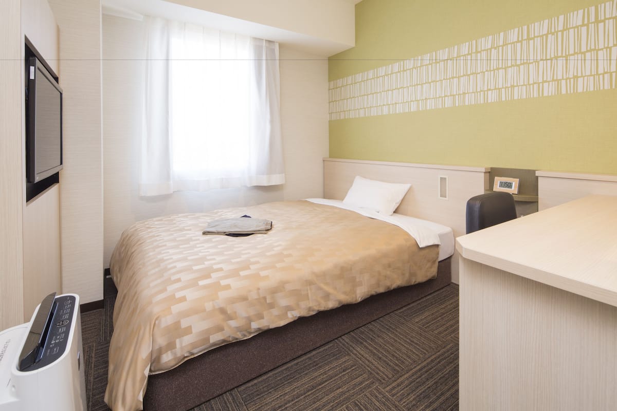 Una habitación de hotel con cama, escritorio, televisor y ventana con cortinas.