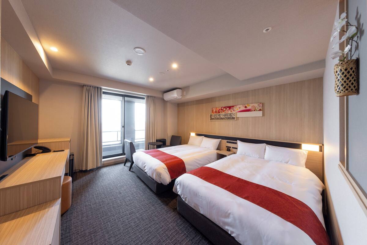 Habitación de hotel con dos camas, un escritorio, una silla, un televisor y una ventana con cortinas.
