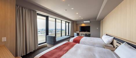 A hotel room with two beds, a desk, a TV, and a large window offering a city view.
