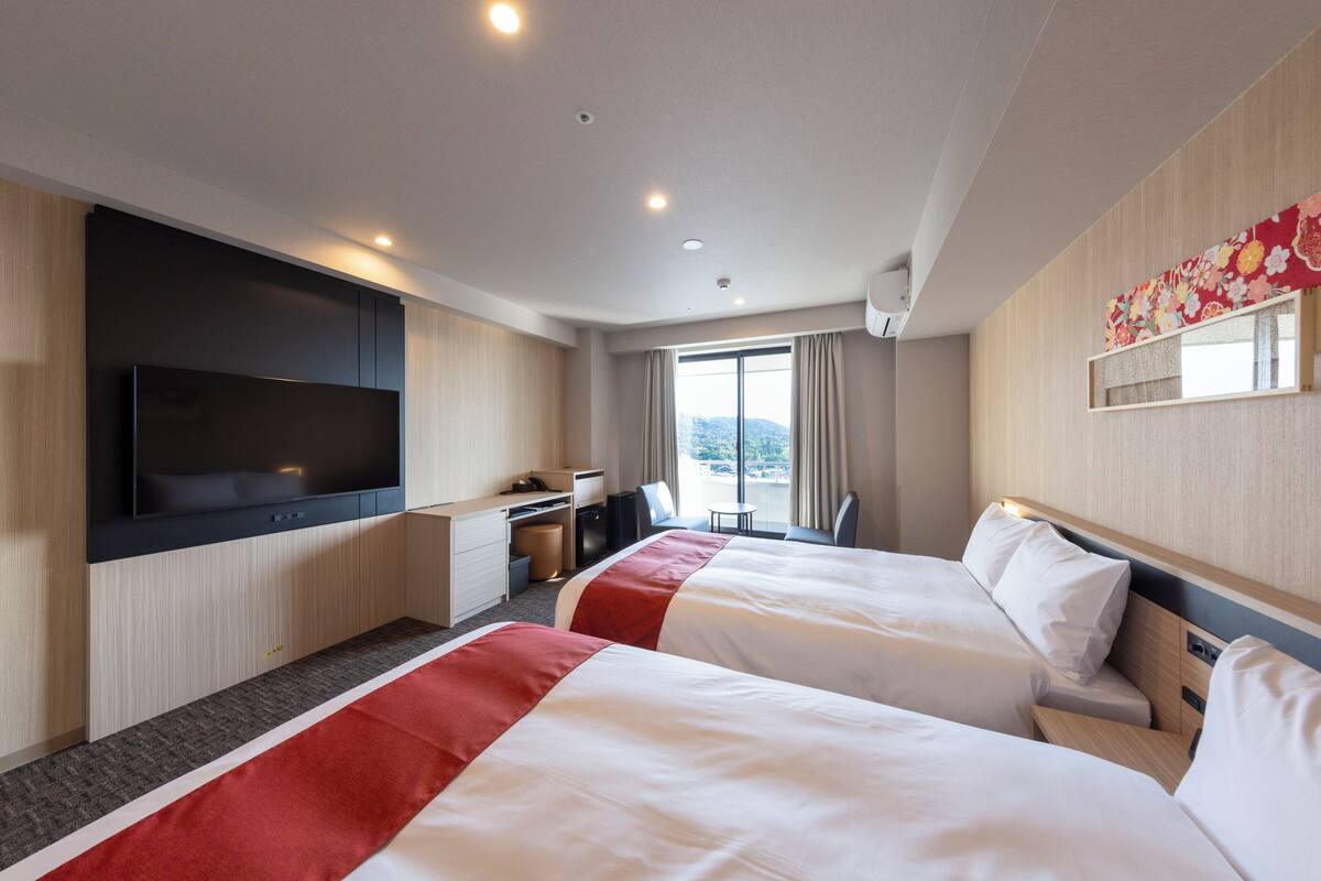 Habitación de hotel con dos camas, televisión, escritorio y un ventanal grande.