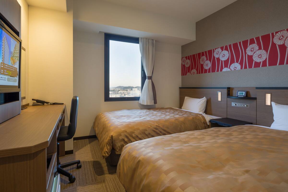Habitación de hotel con dos camas, un escritorio, una silla, una ventana con vistas, un reloj y un panel decorativo en la pared.