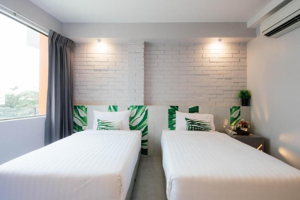 Habitación de hotel con dos camas, ropa de cama blanca, almohadas con estampado de hojas verdes, una cortina gris y una pared con diseño de ladrillo.