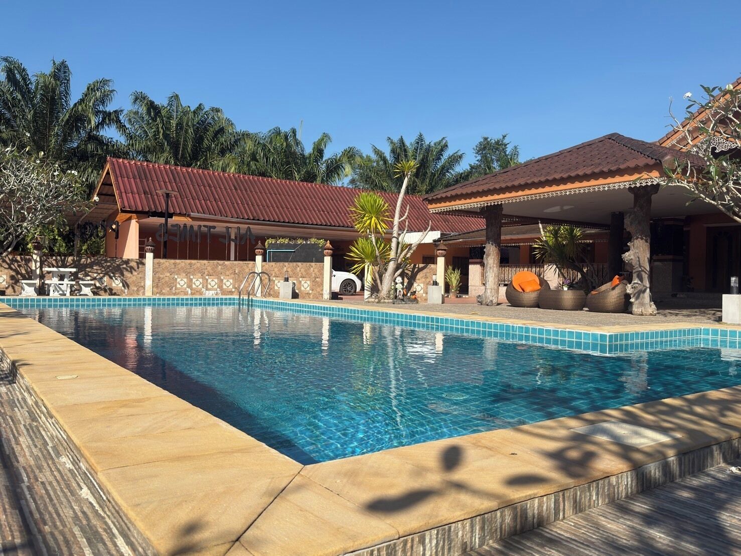 A swimming pool with a tiled edge, a covered patio area, and a building with a red roof.