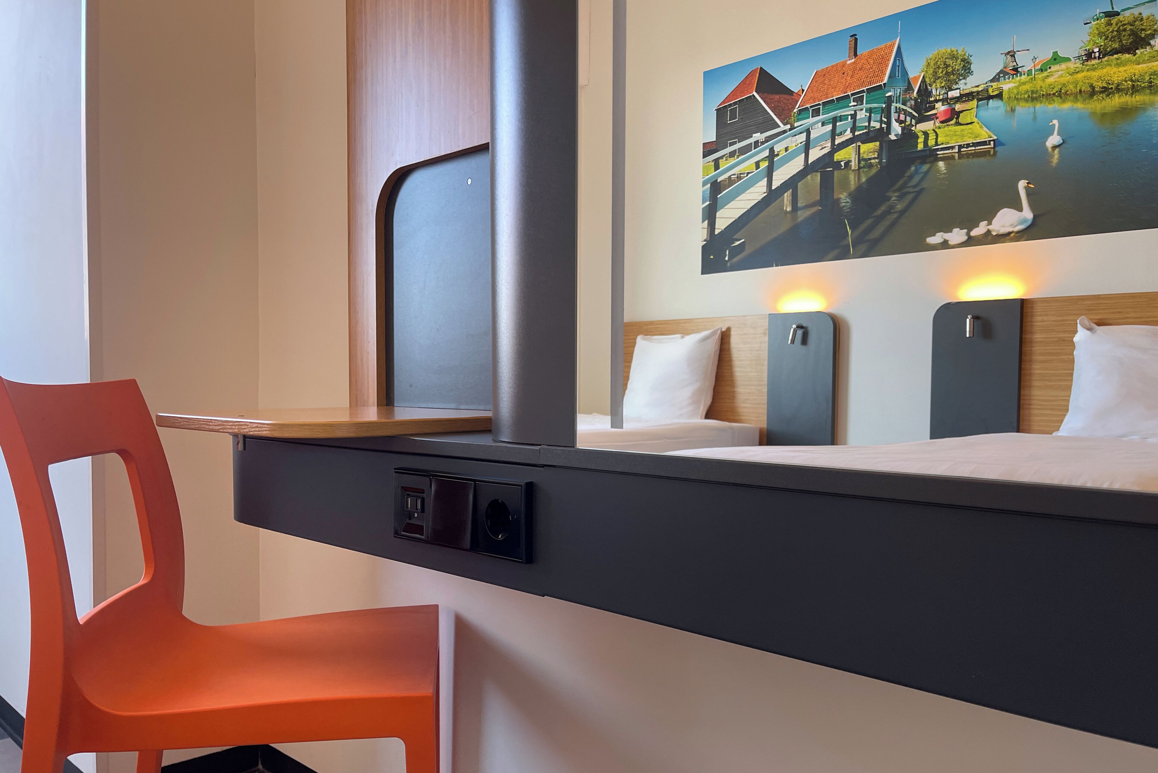 A hotel room with a bed, a desk with a chair, a window, and a painting of a village scene.