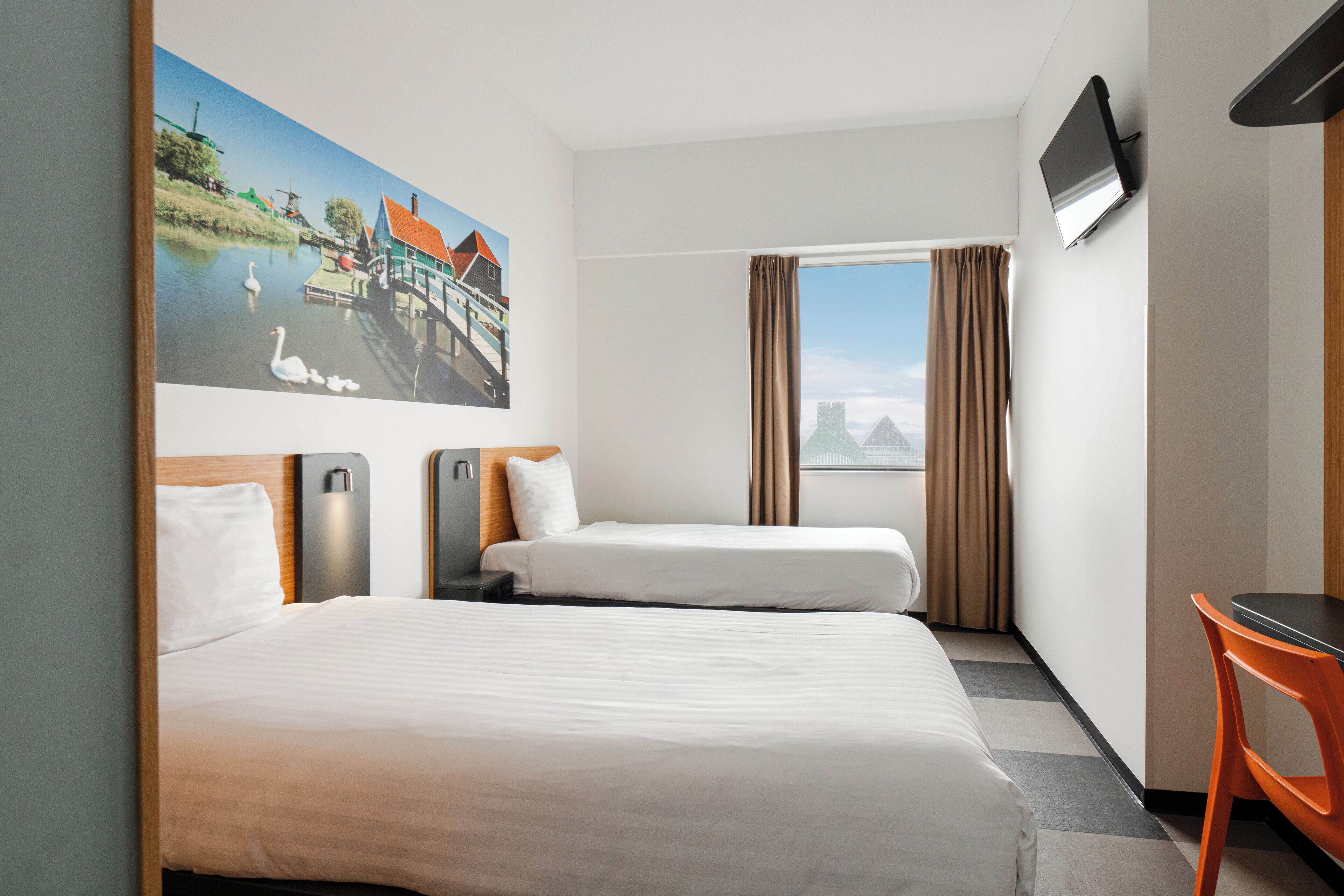 Ein Hotelzimmer mit einem großen Bett, einem kleineren Bett, einem Schreibtisch, einem Sessel, einem Fernseher und einem Landschaftsbild.