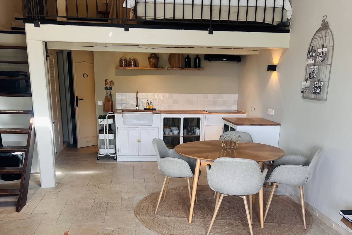 A spacious living area with a high ceiling, a dining table with four chairs, a kitchen with white cabinets, and a staircase leading to an upper level.