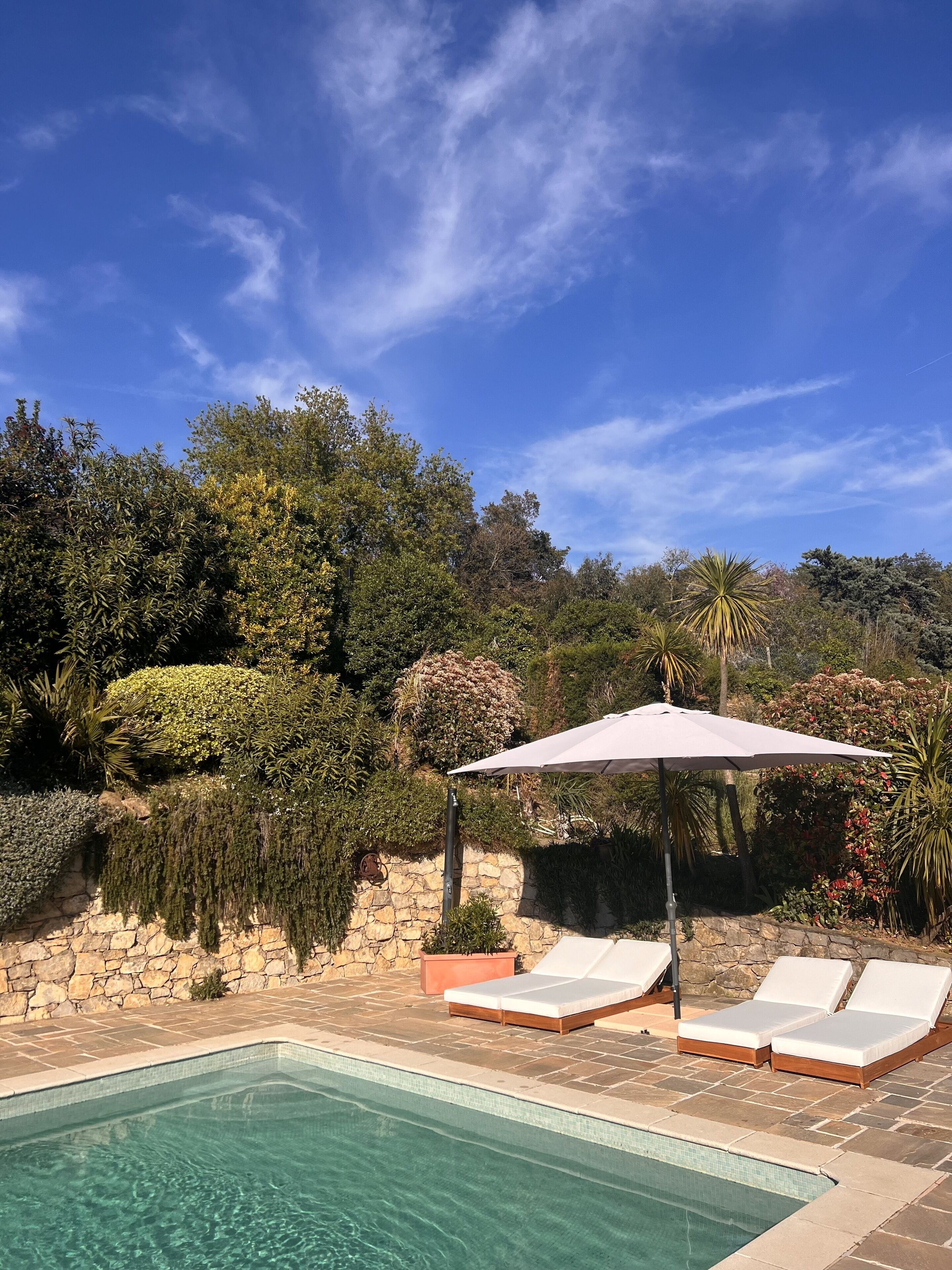 Ein Swimmingpool mit Liegestühlen und Sonnenschirm, umgeben von üppiger Vegetation und einer Steinmauer.
