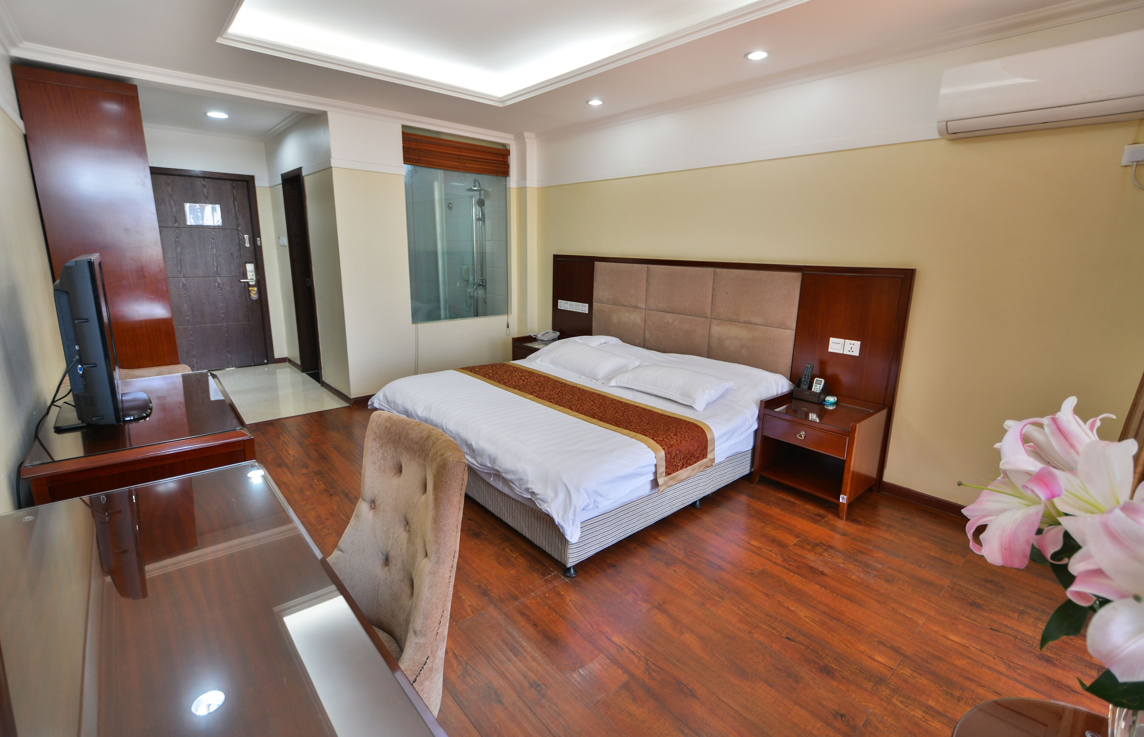 Een hotelkamer met een houten bureau, een bed met een bruin hoofdeinde, een nachtkastje en een douchecabine van glas.
