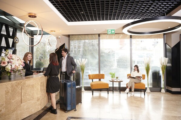A hotel reception area with a modern design, including a reception desk, a seating area with a table and chairs, and a large window providing a view of the city.