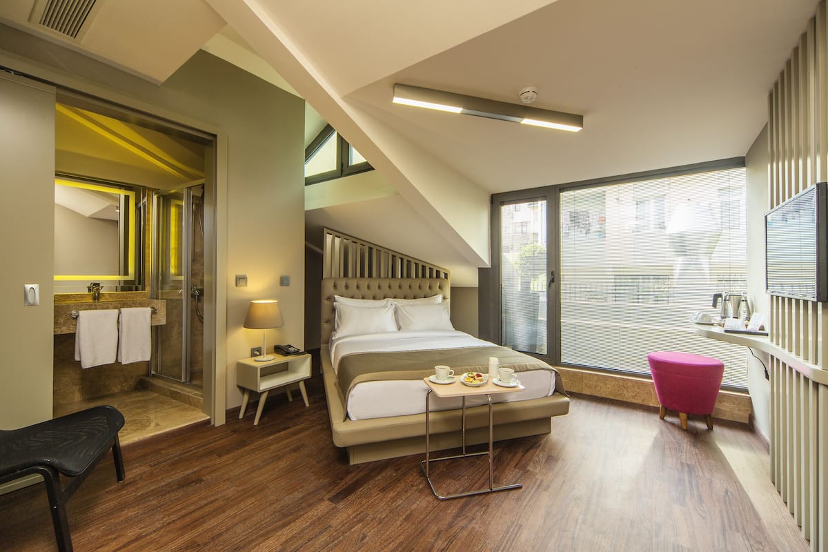 A modern hotel room with a large bed, a small table with breakfast items, a shower area, and a TV on the wall.