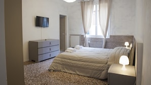 Un dormitorio con cama, cómoda, televisor, mesita de noche con lámpara y ventana con cortinas.