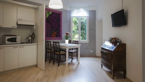 Cocina compacta con armarios blancos, microondas, mesa de comedor con sillas, ventana grande con vistas a zonas verdes y televisor montado en la pared.