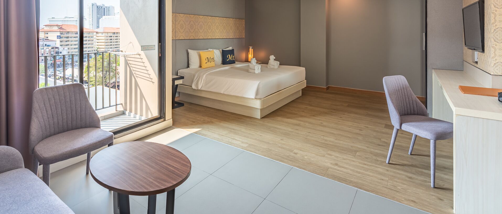 A modern hotel room with a bed, two chairs, a small table, a TV, and a balcony with a city view.