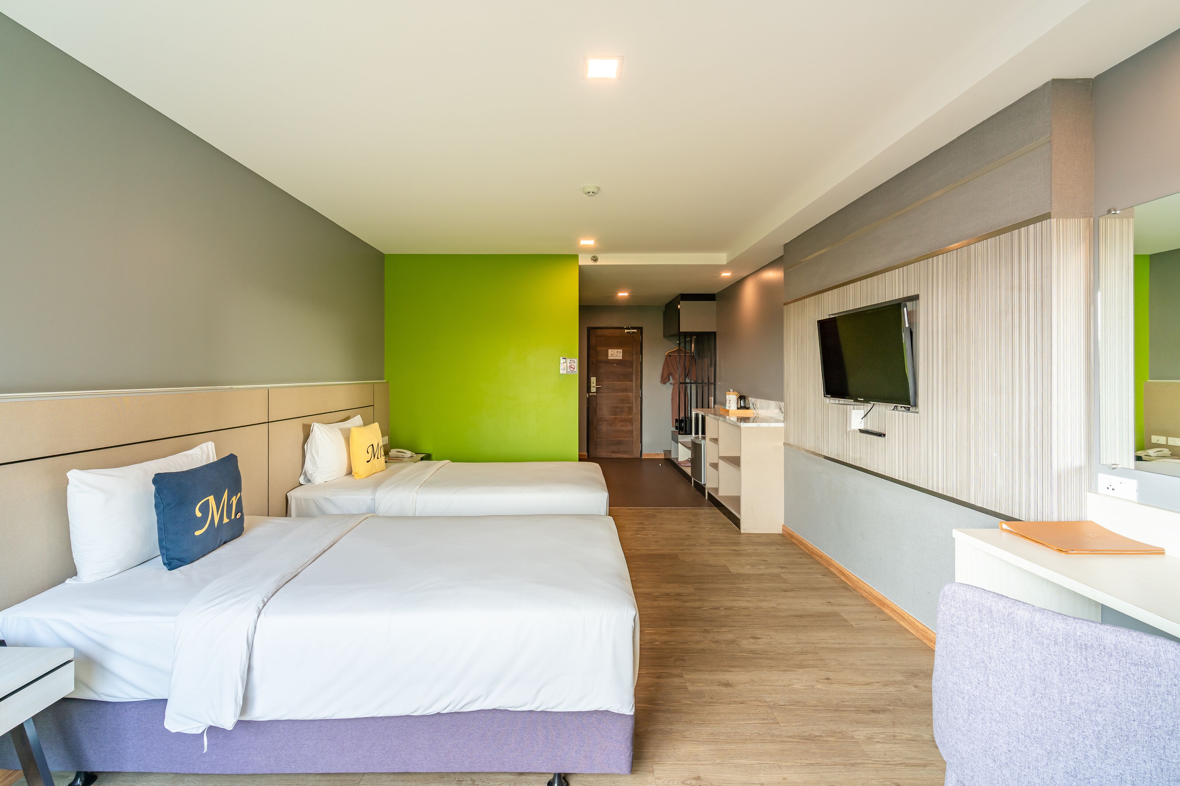 A hotel room with two beds, a desk, a TV, and a bathroom in the background.