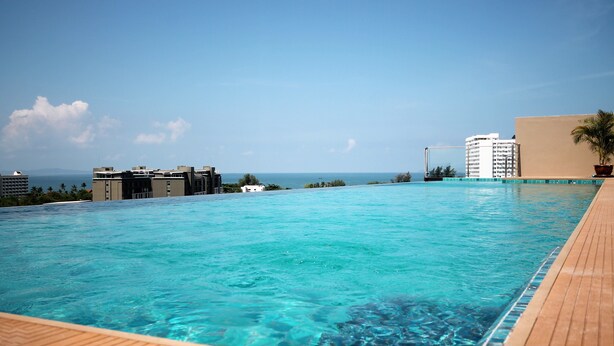 Une piscine rectangulaire à débordement, avec une eau d’un bleu cristallin, une terrasse en bois et une vue sur l’océan et les bâtiments situés au loin.