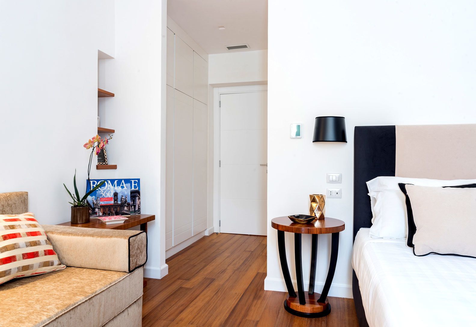Ein modernes Hotelzimmer mit Holzboden, einer Couch mit gemustertem Kissen, einem kleinen runden Tisch mit einer Dekoration und einem Bett mit weißer Bettwäsche, sowie einer Wandlampe.
