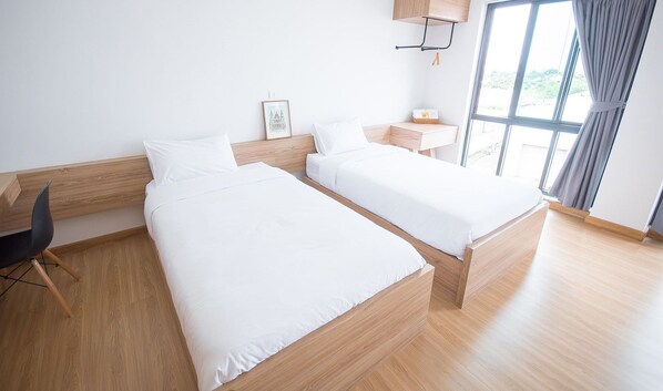 Habitación de hotel con dos camas individuales, piso de madera, un escritorio pequeño con silla, una ventana con cortinas y un estante en la pared con un cuadro.