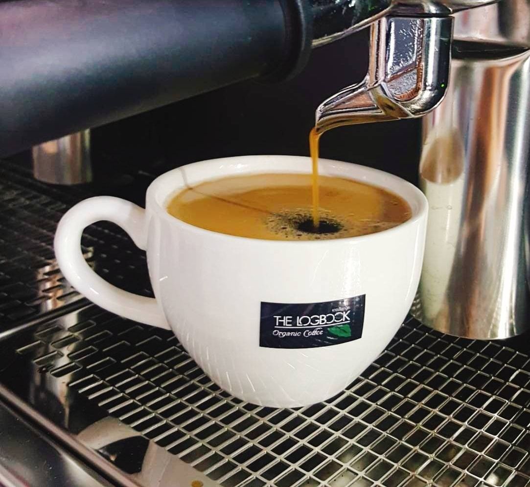 Uma chávena de café branca com um logótipo, a ser enchida com café por uma máquina.