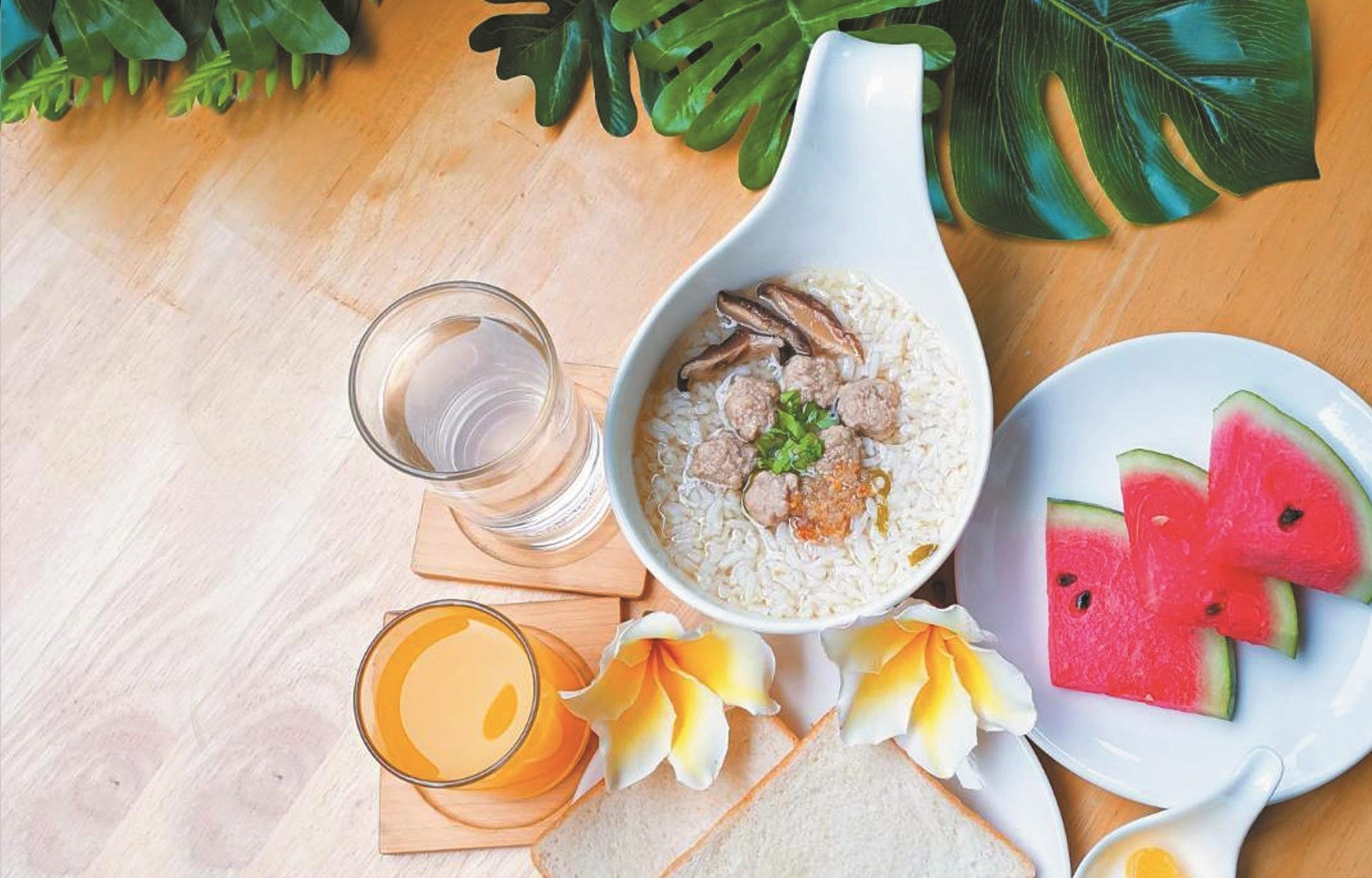 A meal setup with a bowl of rice, meatballs, and mushrooms, a glass of orange juice, slices of watermelon, and a glass of water on a wooden table with a tropical plant in the background.