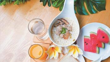 A meal setup with a bowl of rice, meatballs, and mushrooms, a glass of orange juice, slices of watermelon, and a glass of water on a wooden table with a tropical plant in the background.