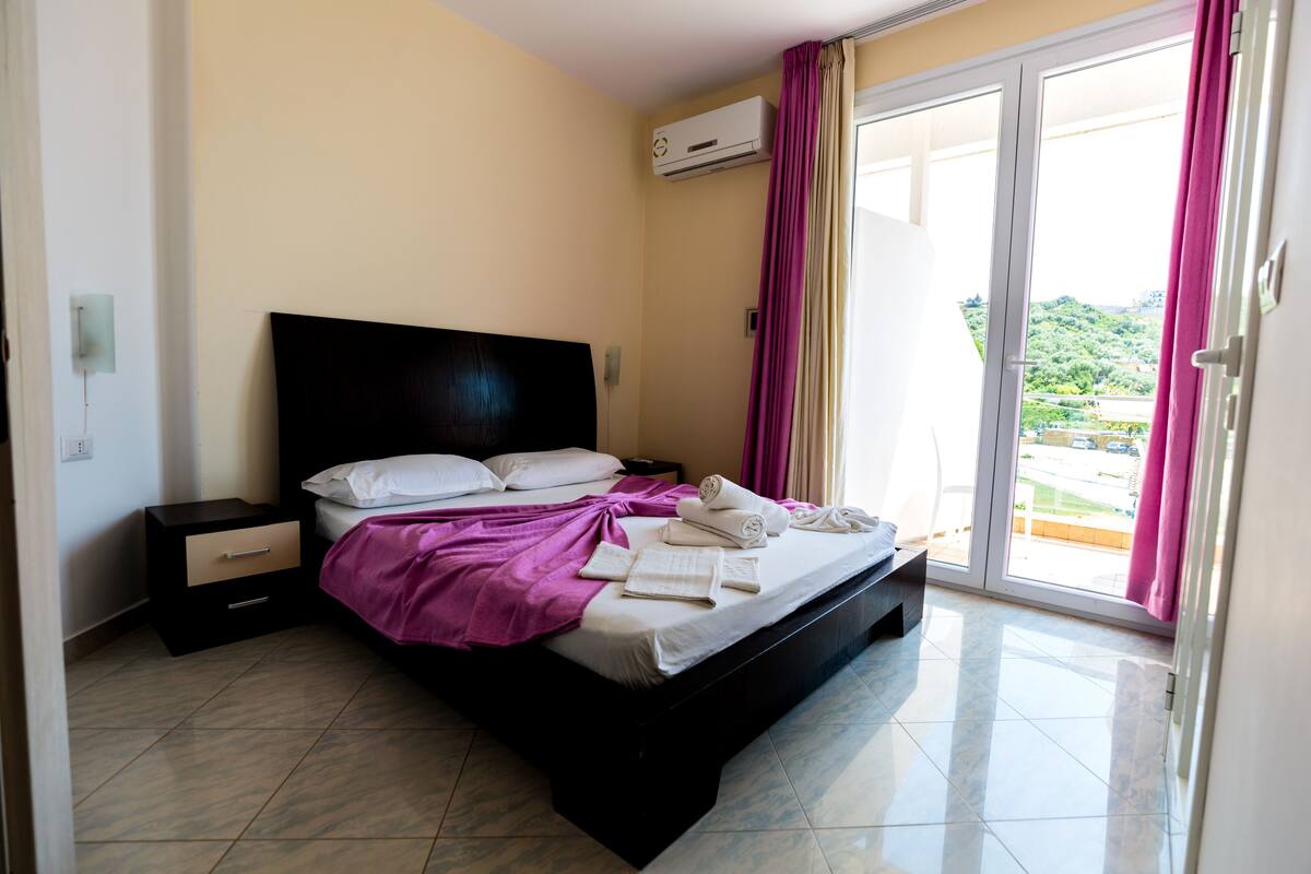 A bedroom with a bed, a nightstand, a window with curtains, and an air conditioning unit.
